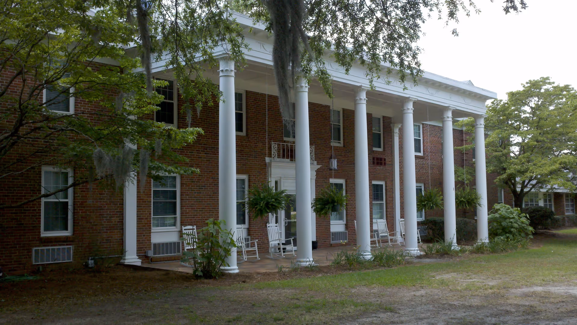 Front exterior view of a brick building with white columns supporting a covered porch. The porch has several white rocking chairs and hanging plants. Trees and greenery surround the building.