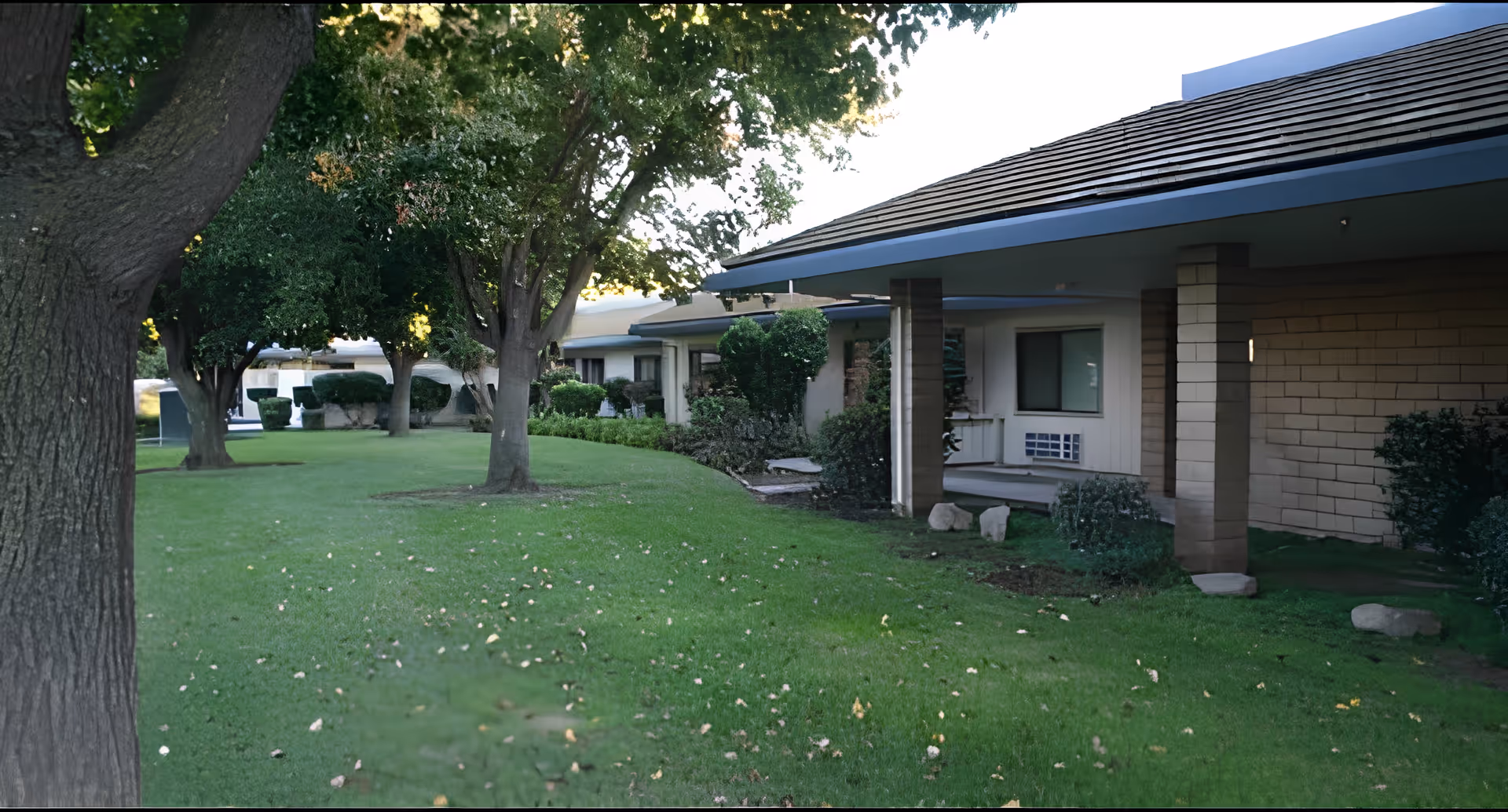 View of a single-story assisted living facility building with a covered walkway, surrounded by green grass, trees, and shrubs under natural daylight.