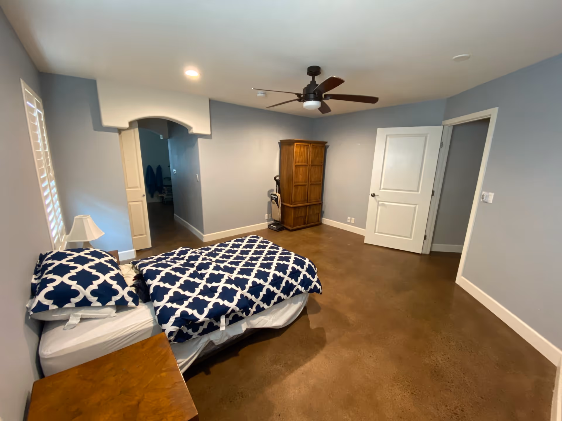 Simple bedroom with bed, navy patterned bedding, dresser, lamp, and ceiling fan.