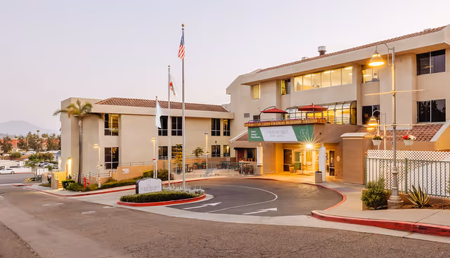 Exterior view of Country Hills Post Acute facility at dusk, showing a multi-story building with beige walls and red-tiled roofs, a circular driveway with a landscaped island featuring three flagpoles with flags, and street lamps illuminating the entrance area.