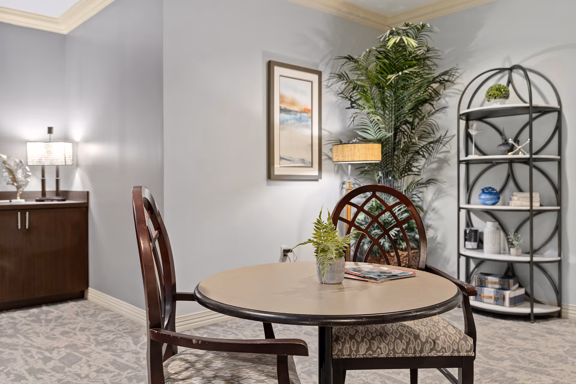 A cozy interior room with a round table and two wooden chairs with patterned cushions. On the table is a small potted plant and some magazines. In the background, there is a tall green plant, a framed abstract painting on the wall, a black metal shelf with decorative items, and a wooden cabinet with a lamp on top. The room has light gray walls and carpeted flooring.