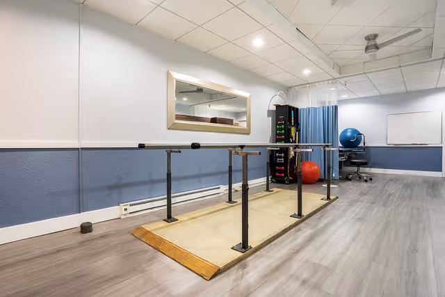 A rehabilitation or physical therapy room with parallel bars for walking exercises, a large mirror on the wall, exercise equipment including a blue exercise ball and a red ball, a chair, and a whiteboard in the background. The room has light blue and white walls and wood-style flooring.