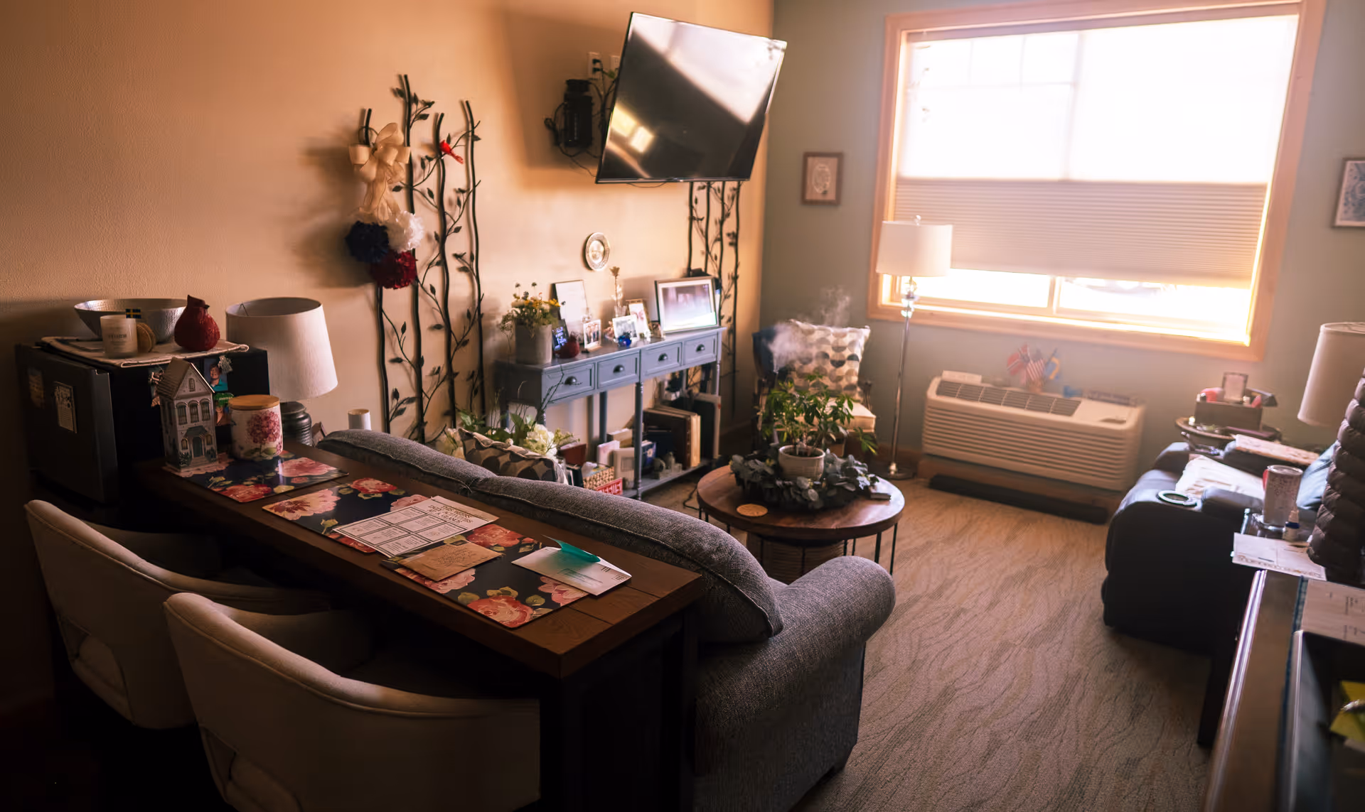 A cozy living room with a gray couch facing a wall-mounted TV. There is a wooden console table behind the couch with two white chairs. The room has a round coffee table with plants, a floor lamp near a window with blinds, and an air conditioning unit below the window. Decorative items and framed photos are on a side table against the wall.