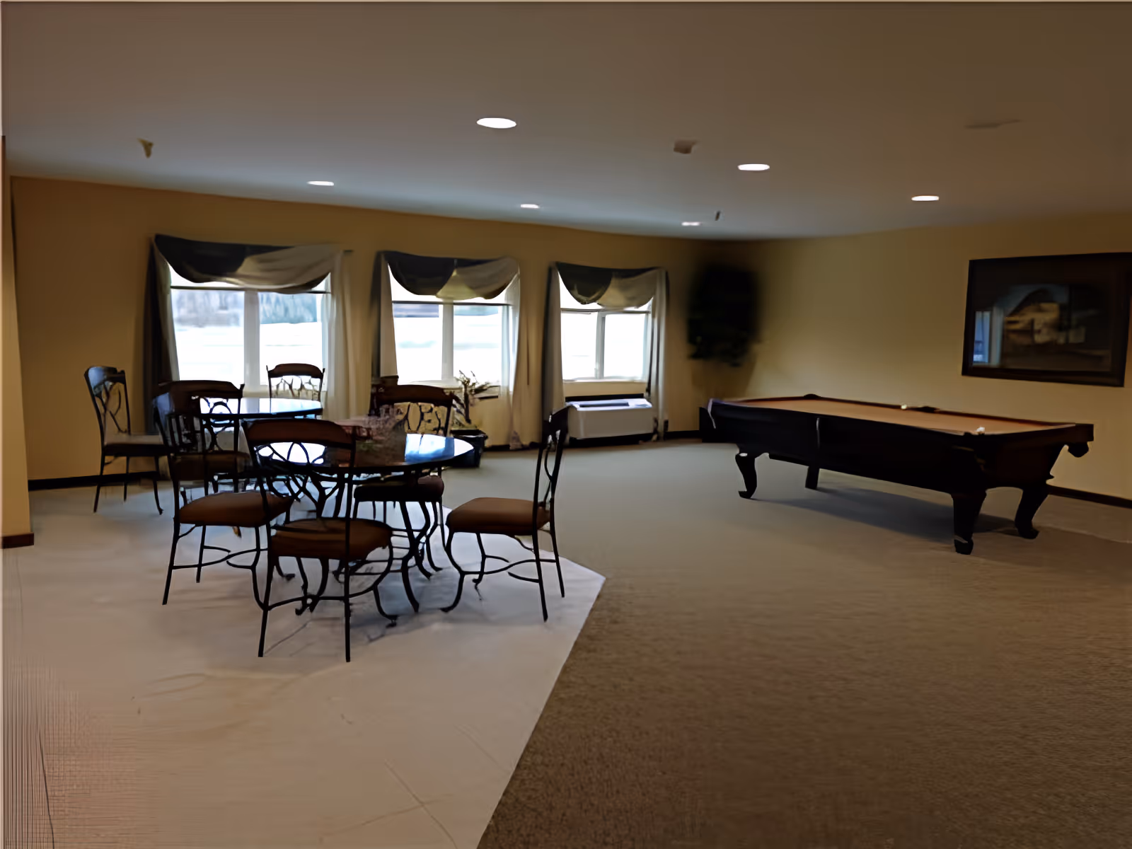 A spacious room with a pool table on the right side and a seating area with round tables and chairs on the left side near three large windows with curtains. The walls are painted light yellow and there is a framed picture hanging on the wall above the pool table.