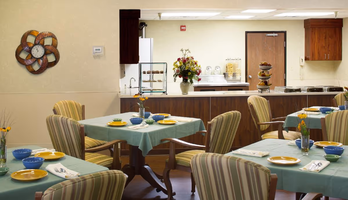 Dining area with tables covered in green tablecloths set with yellow plates, blue bowls, and silverware. Striped upholstered chairs surround the tables. In the background, there is a kitchen counter with a flower vase, a tiered tray with fruit, and kitchen appliances. A decorative wall clock is mounted on the left wall.