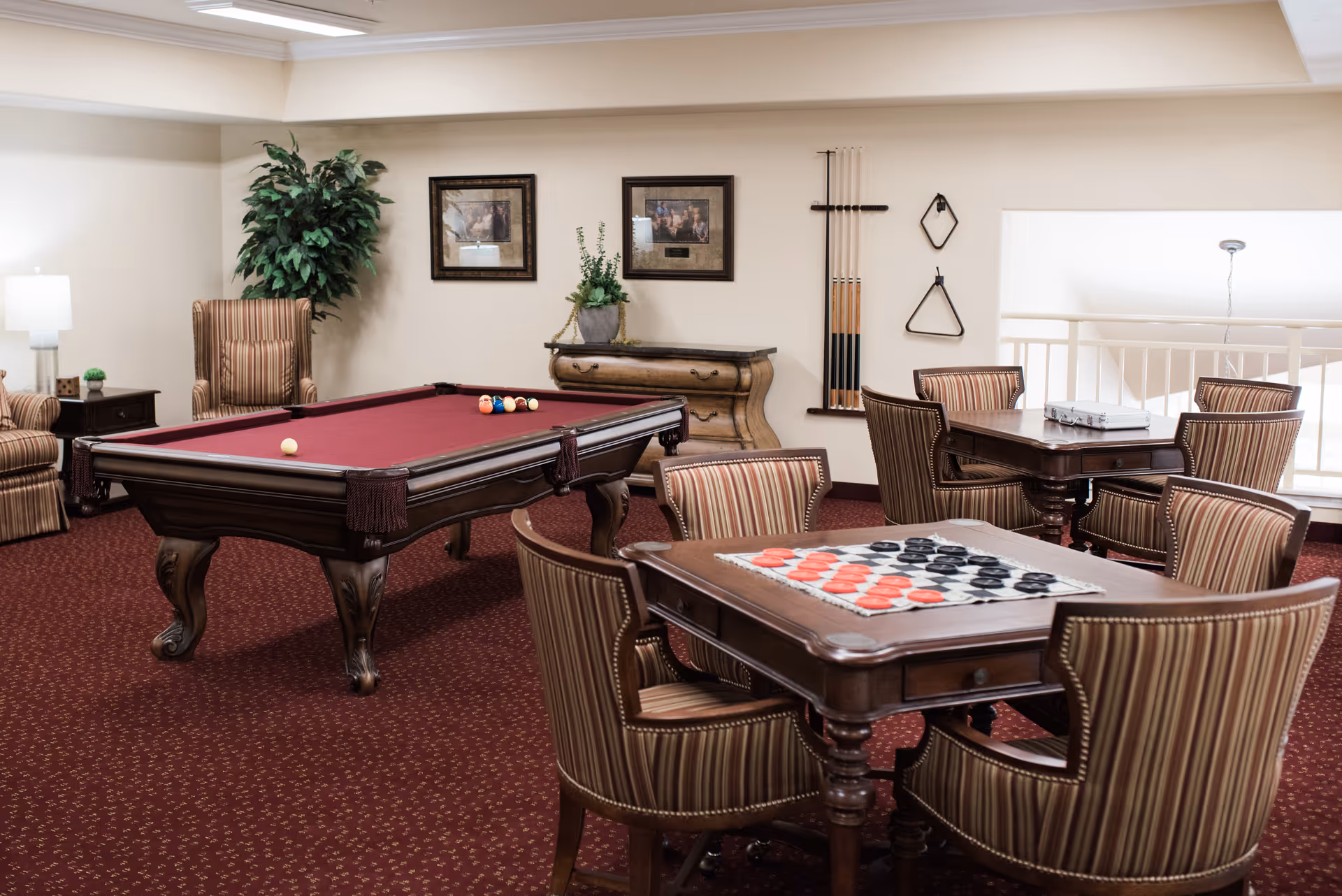 A cozy game room with a burgundy pool table, a table with a checkers board, several striped upholstered chairs, a side table with a lamp, a potted plant, and framed pictures on the wall. Pool cues and triangle racks are mounted on the wall.