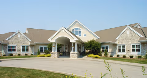 Front exterior view of a single-story senior living facility building with beige siding and stone accents, a covered entrance with columns, and a circular driveway surrounded by grass and landscaping.
