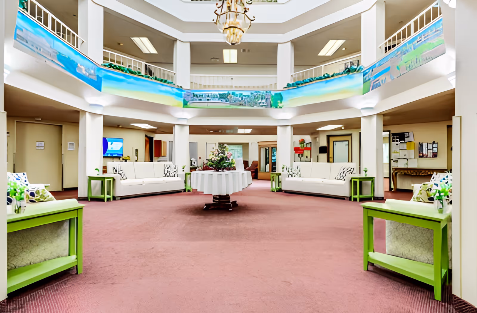 Bright atrium-style lobby with sofas, green side tables and a central table of flowers beneath a two-story balcony.