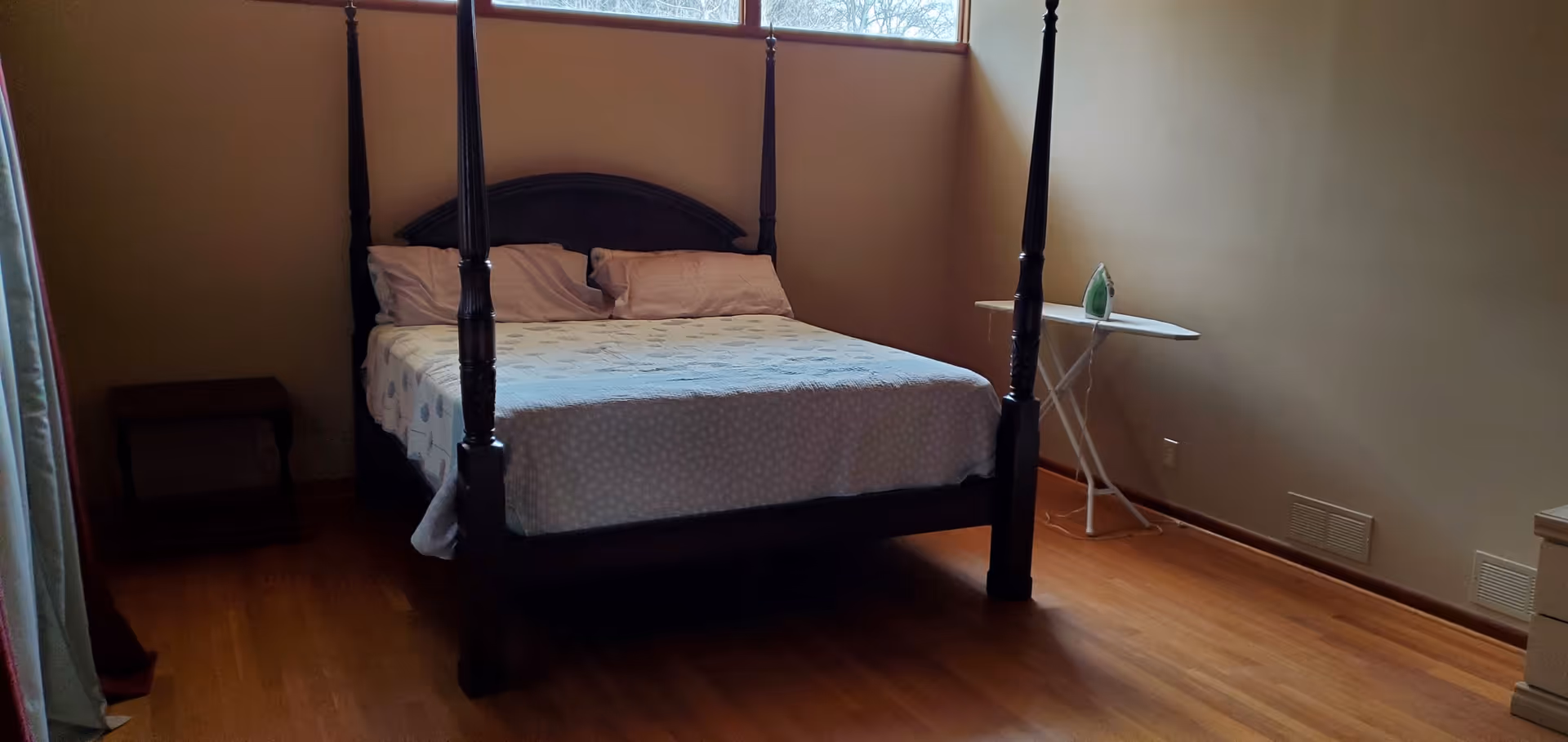 A bedroom with a four-poster bed made with white and patterned bedding, two pillows, a small wooden side table on the left, and an ironing board with an iron on the right. The room has wooden flooring and a high window letting in natural light.