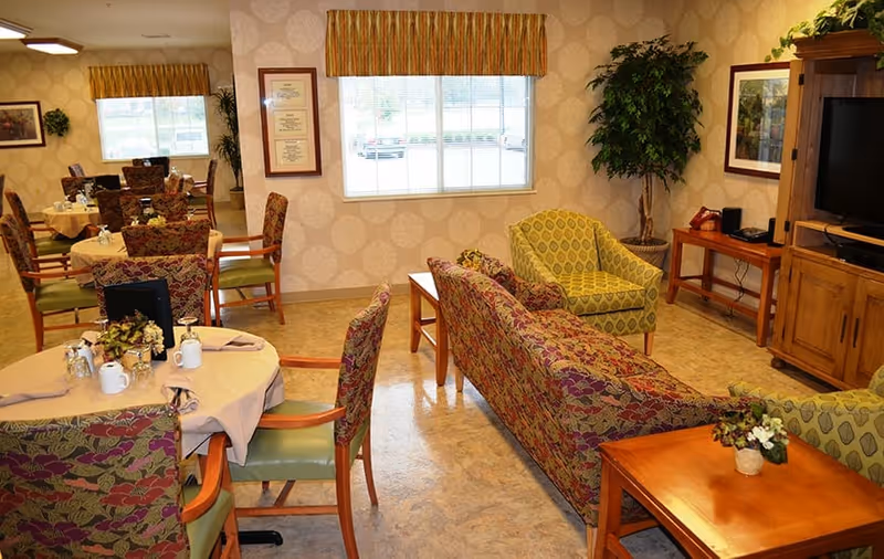 A cozy common area in a senior living facility featuring several round tables with floral-patterned chairs, set with cups and napkins. There is a patterned sofa and armchairs arranged around a wooden coffee table. A large window with striped valance lets in natural light, and the walls are decorated with framed pictures and certificates. A wooden entertainment center with a TV and a potted plant are also visible.