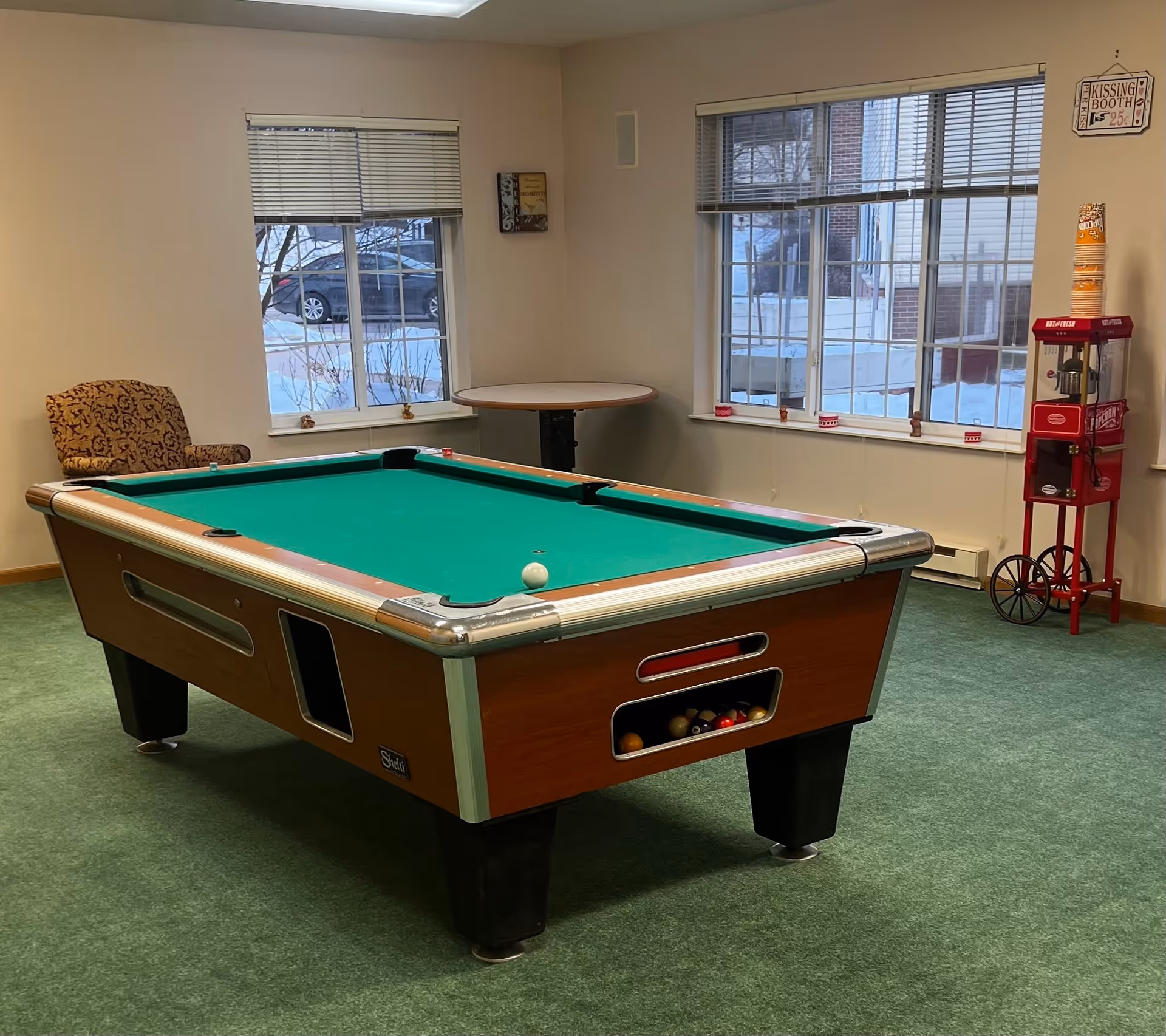 A recreational room with a pool table in the center, a patterned armchair near a window, a round table against the wall, and a red popcorn machine with stacked popcorn containers on top. The room has green carpet and two large windows showing a snowy outdoor scene.