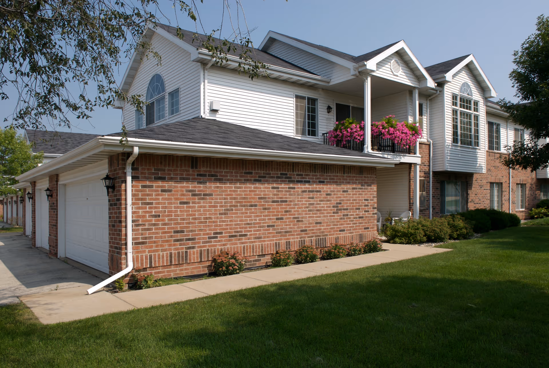 Exterior view of a two-story brick and white-siding apartment building with attached garages, a balcony with pink flowers, and a green lawn.