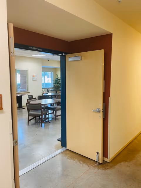Open doorway into a communal dining/activity room with round tables and chairs.