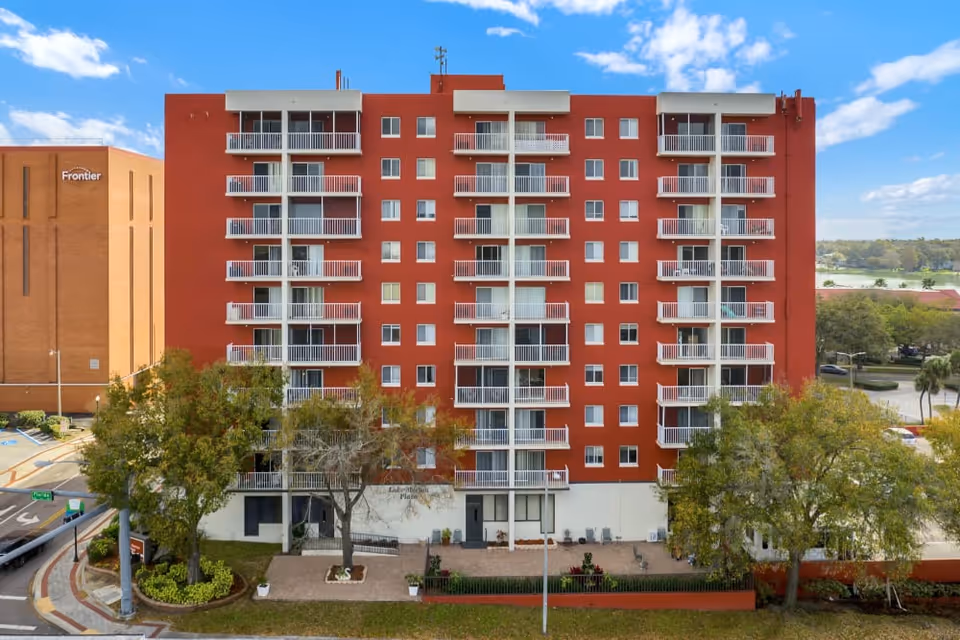 Exterior view of a multi-story red brick building with white balconies, identified as Lake Morton Plaza. The building is surrounded by trees and landscaping, with a clear blue sky in the background. A neighboring building with the Frontier logo is visible on the left side.