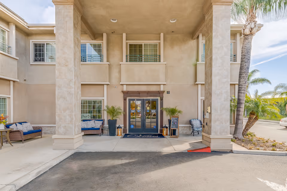 Entrance of Villas at San Bernardino facility showing double glass doors under a covered area with two large columns. There are outdoor seating areas with cushioned chairs and small tables on either side of the entrance, potted plants, and a welcome sign near the door. Palm trees and a clear sky are visible in the background.