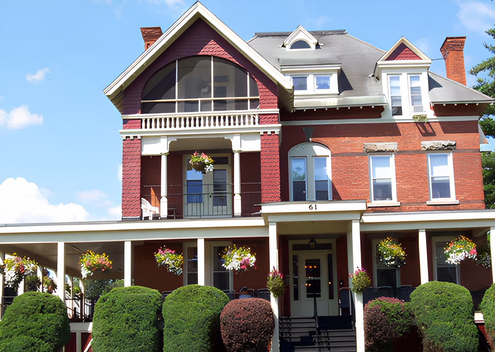Front exterior of a large historic red-brick three-story house with a wraparound porch, hanging flower baskets, and trimmed shrubs.