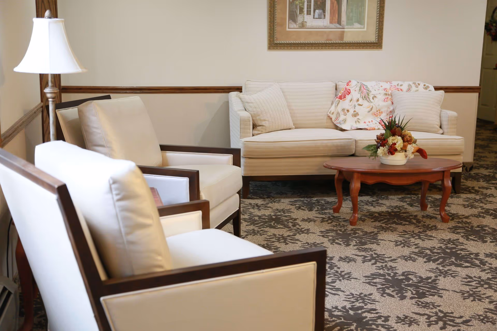 A cozy living room area with a beige sofa adorned with two cushions and a floral throw blanket. In front of the sofa is a wooden coffee table with a floral arrangement. Two cream-colored armchairs with wooden frames are positioned nearby. A floor lamp stands in the corner, and a framed picture hangs on the wall above the sofa. The carpet has a floral pattern in shades of brown and beige.