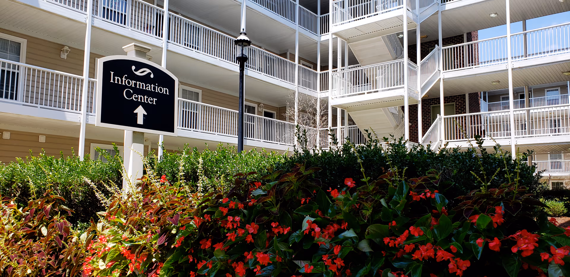 Courtyard view of a multi-story apartment building with white balconies, a sign reading "Information Center," and flowering shrubs in the foreground.
