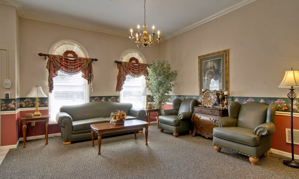 A furnished common living room with green leather sofa and chairs, a wooden coffee table, lamps, chandelier, and decorative accents by two arched windows.