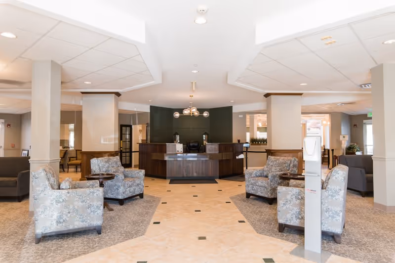A spacious and well-lit senior living facility lobby with a central reception desk in dark wood, surrounded by comfortable floral-patterned armchairs arranged in small seating areas. The floor is tiled with a combination of beige tiles and carpeted sections. The ceiling has recessed lighting and a modern chandelier above the reception desk. A hand sanitizer dispenser is visible in the foreground.