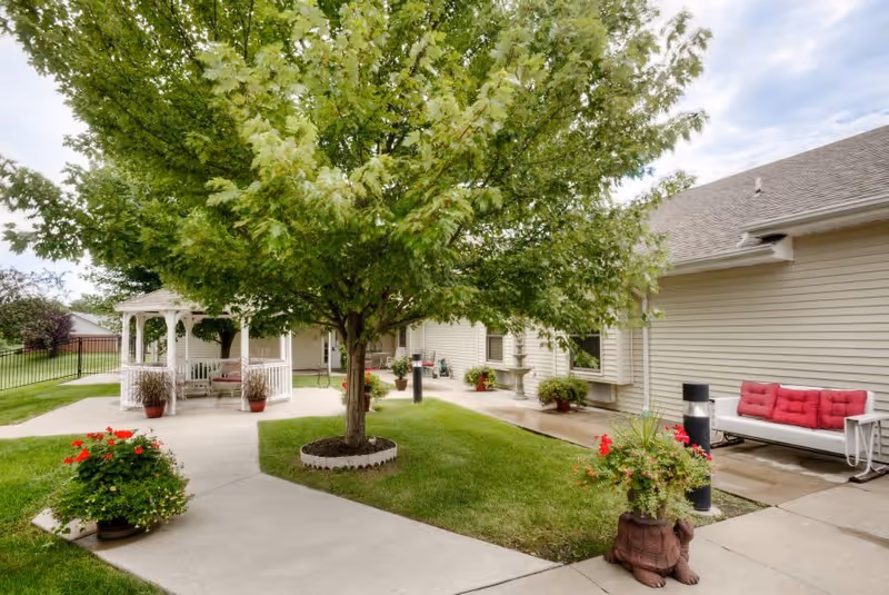Outdoor courtyard area with a large leafy tree in the center surrounded by a concrete walkway. There is a white gazebo with seating on the left side and a white bench with red cushions on the right side. Various potted plants and flowers are placed around the courtyard, and the building exterior is beige with windows.