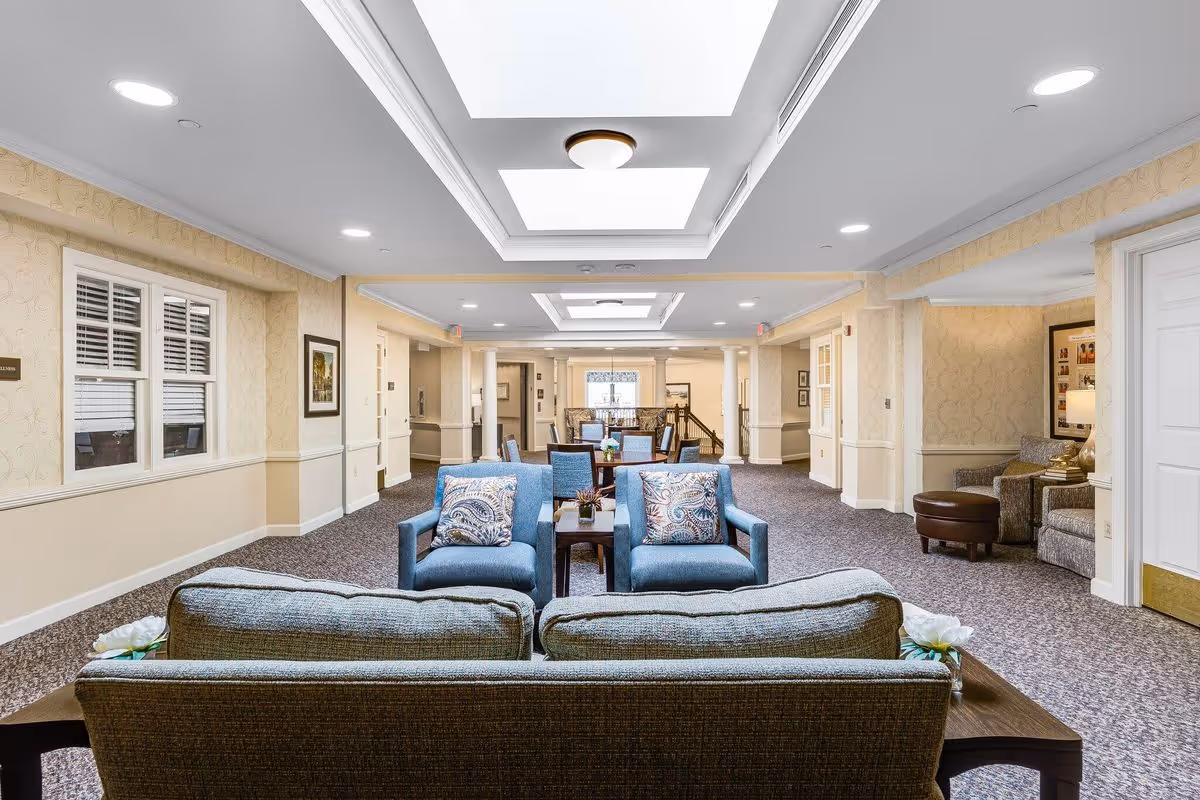 A spacious and well-lit common area in a senior living facility featuring comfortable seating including a sofa and two armchairs with patterned cushions. The room has beige walls with white trim, carpeted floors, recessed lighting, and skylights. There are tables with decorative plants and framed artwork on the walls.