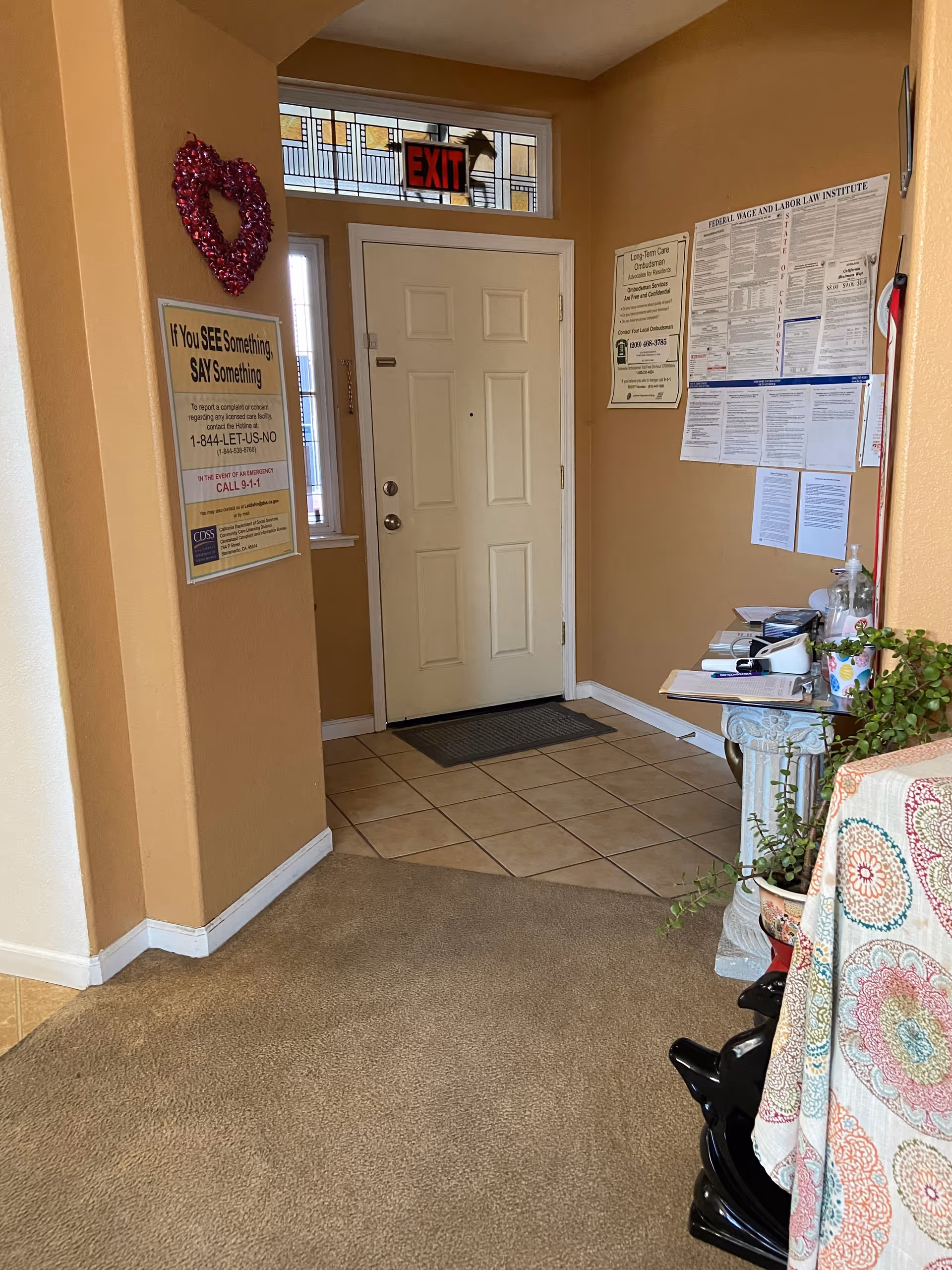 Interior view of an entryway with a closed white door, a small window above the door with an illuminated red EXIT sign, and beige walls. There are informational posters on the walls, a small table with papers and plants on the right, and a carpeted floor transitioning to tiled flooring near the door.