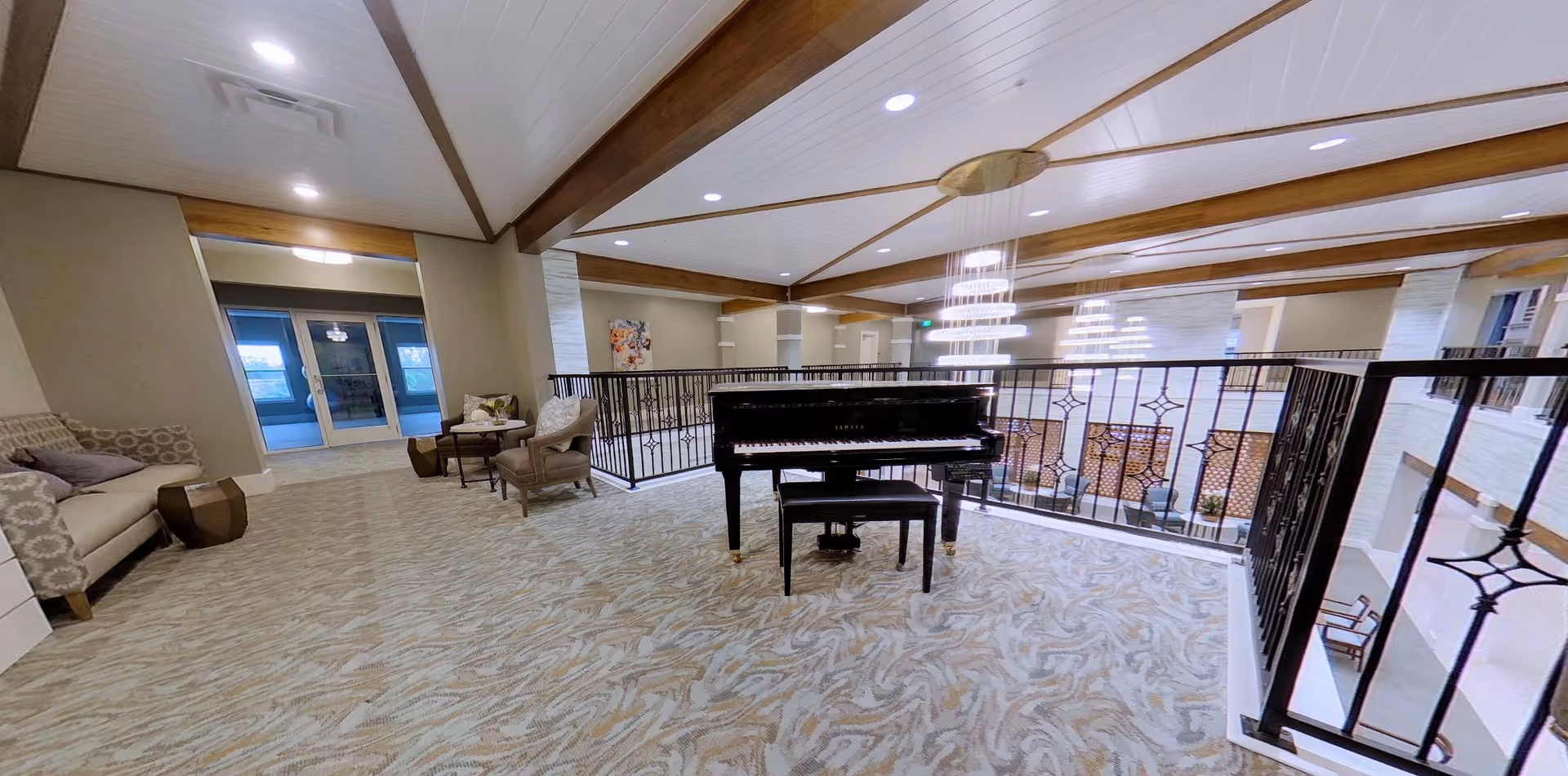 Spacious indoor common area with a grand piano in the center, surrounded by comfortable seating including armchairs and a sofa. The area features a patterned carpet, wooden ceiling beams, and modern lighting fixtures. A railing overlooks a lower level with additional seating and large windows allowing natural light.