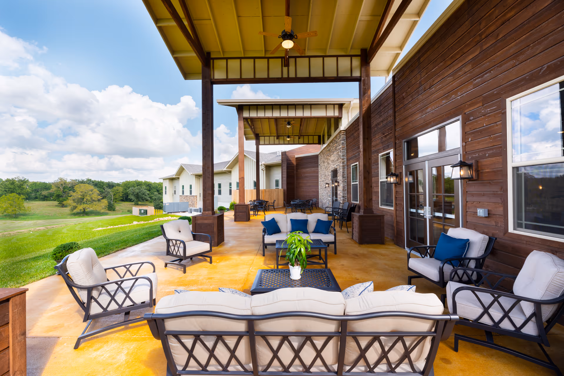 Covered outdoor patio with cushioned seating and tables overlooking a grassy landscape at an assisted living facility.