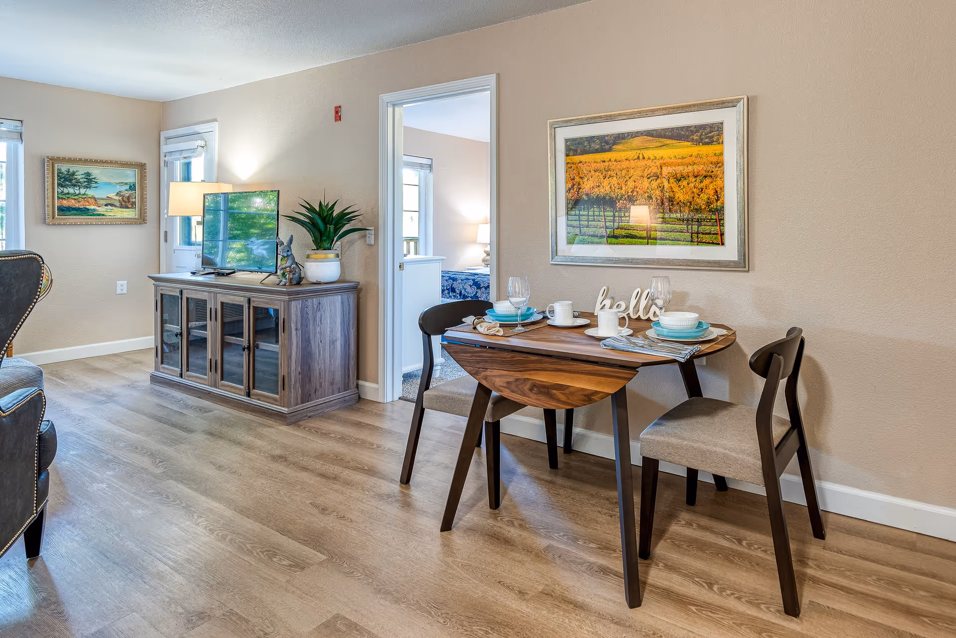 A cozy senior living interior featuring a small wooden dining table set for two with plates, cups, and glasses. The table is accompanied by two cushioned chairs. On the wall above the table is a framed picture of a vineyard landscape. To the left, there is a wooden TV stand with a flat-screen TV, a potted plant, and a decorative figurine. The room has light-colored walls and wood flooring, with a doorway leading to a bedroom visible in the background.