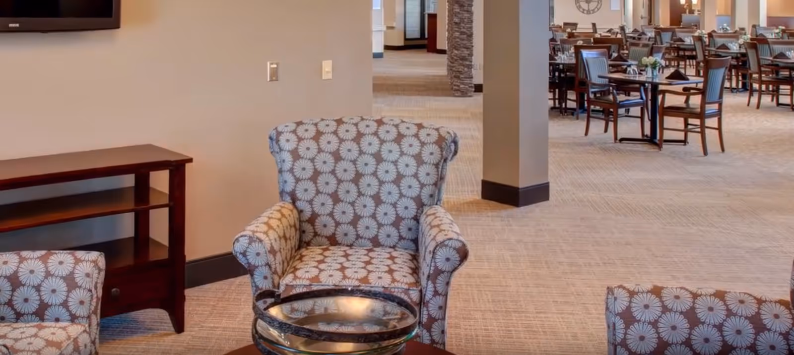 A seating area with patterned armchairs and a small wooden table with a glass bowl on it, adjacent to a dining room with multiple tables and chairs set for meals in a senior living facility.