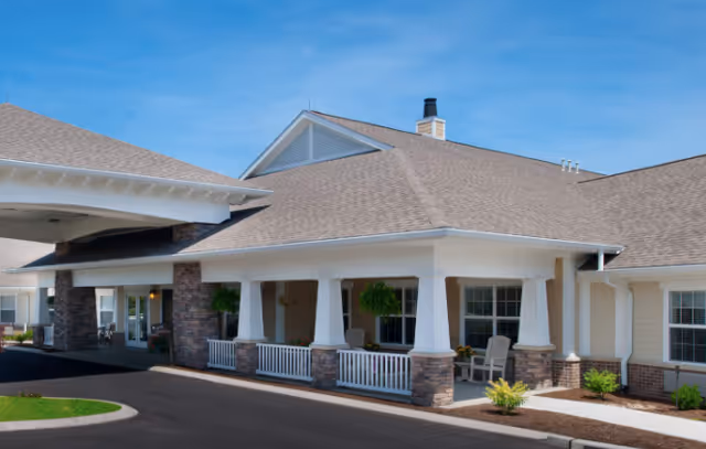 Exterior view of a single-story senior living facility building with a covered entrance, stone pillars, white railings, and a well-maintained driveway under a clear blue sky.