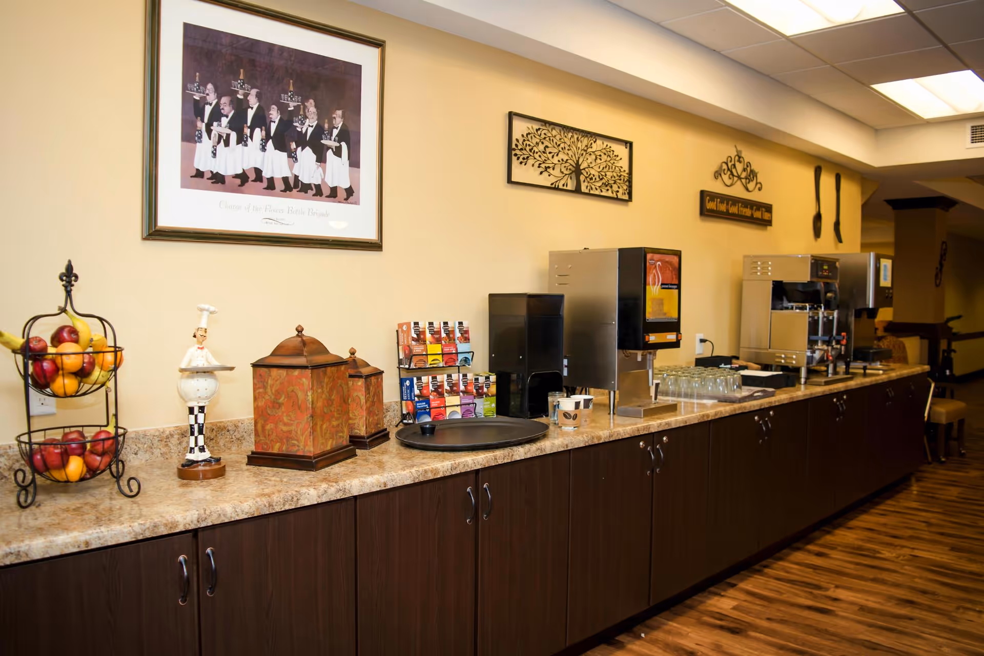 A long countertop with various beverage dispensers, a two-tier fruit basket filled with bananas and apples, decorative containers, and a small chef figurine. Above the counter are framed wall decorations including a painting of waiters and a metal tree artwork. The floor is wooden and the room has a warm, inviting atmosphere.