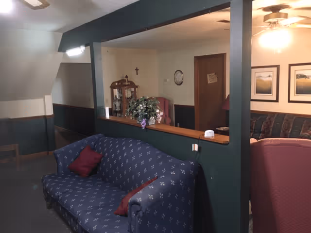 Interior view of a senior living facility showing a blue patterned couch with two red pillows in the foreground. Behind a half wall with a large open window, there is a room with a wooden cabinet, a cross on the wall, framed pictures, and a ceiling fan with a light. The walls are painted green and beige, and there is a hallway visible to the left.