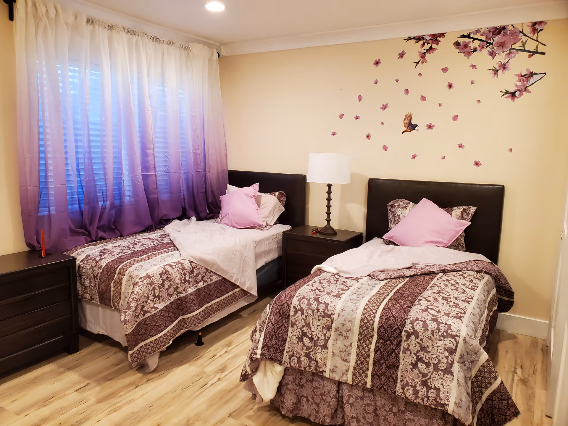 A bedroom with two twin beds featuring patterned bedspreads in shades of purple and white, each with a pink pillow. Between the beds is a dark wooden nightstand with a lamp. The wall behind the beds has decorative stickers of pink flowers and a bird. A window with purple ombre curtains is on the left side of the room, and the floor is light wood.