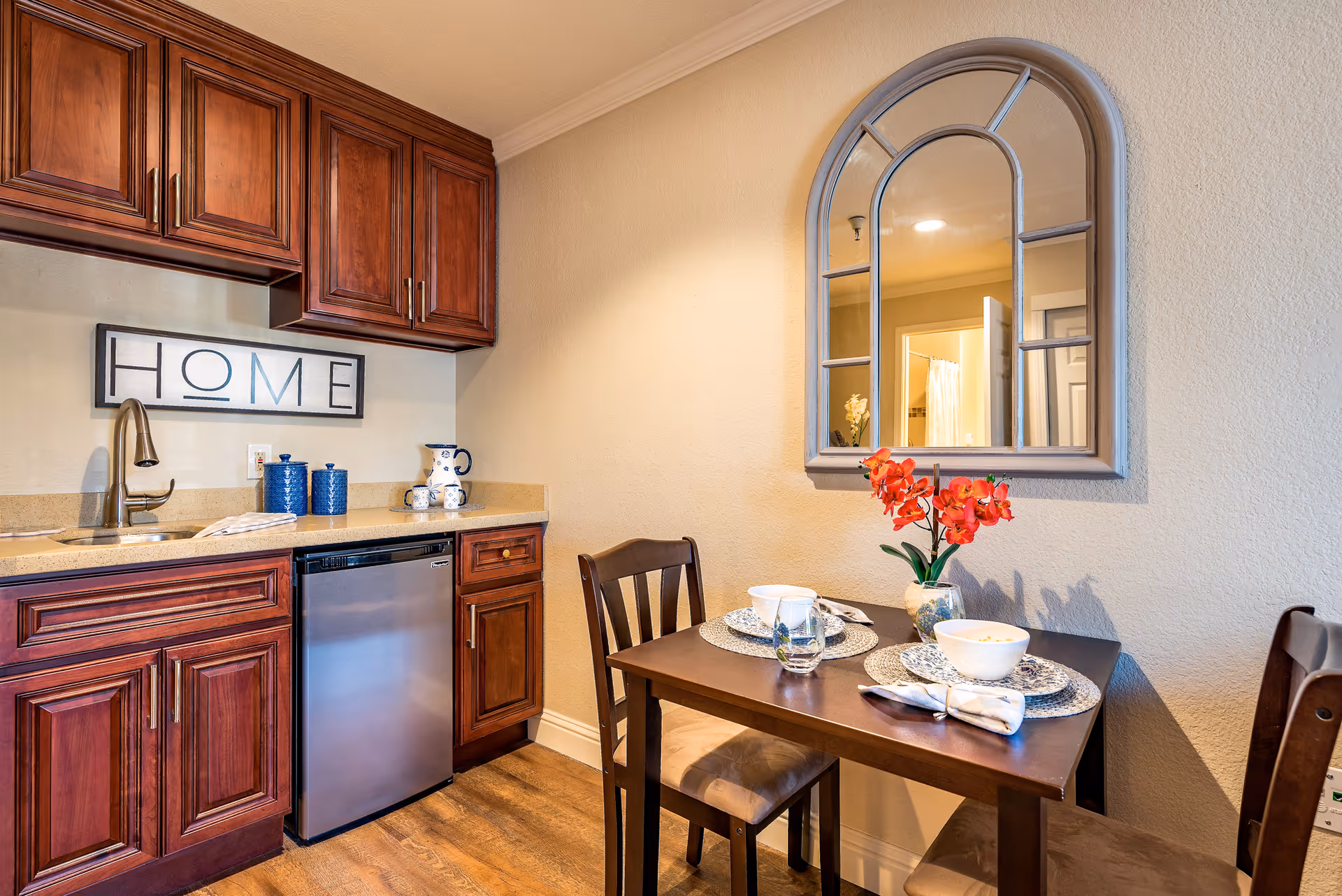 A small dining area and kitchenette with wooden cabinets, a stainless steel mini fridge, a sink, and a countertop. The dining table is set for two with plates, bowls, glasses, napkins, and a vase with red flowers. A decorative mirror hangs on the wall above the table, and a framed sign that reads 'HOME' is mounted above the sink.