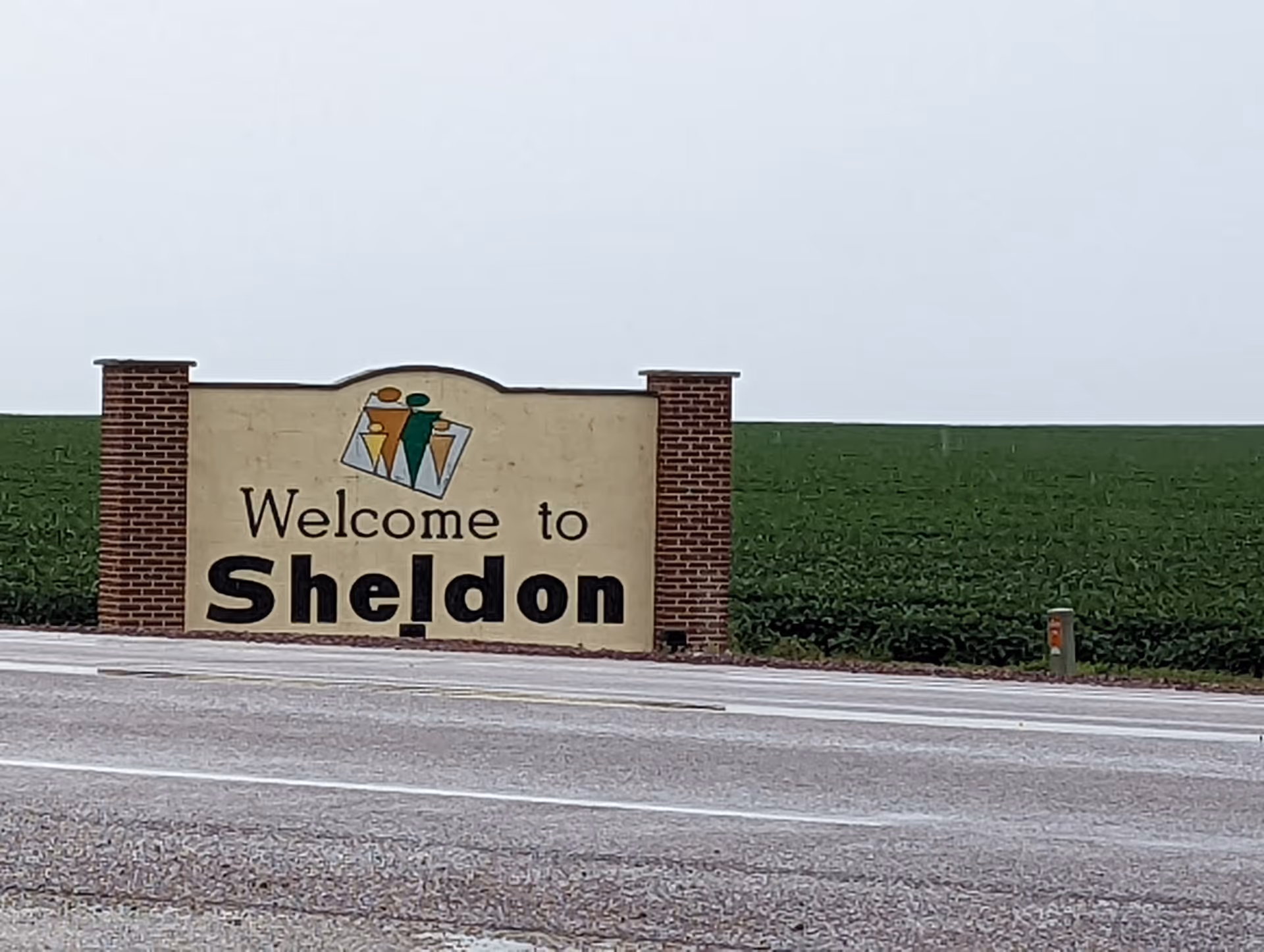 A roadside welcome sign for the town of Sheldon, featuring a beige background with brick pillars on each side and a graphic of three stylized human figures above the text.