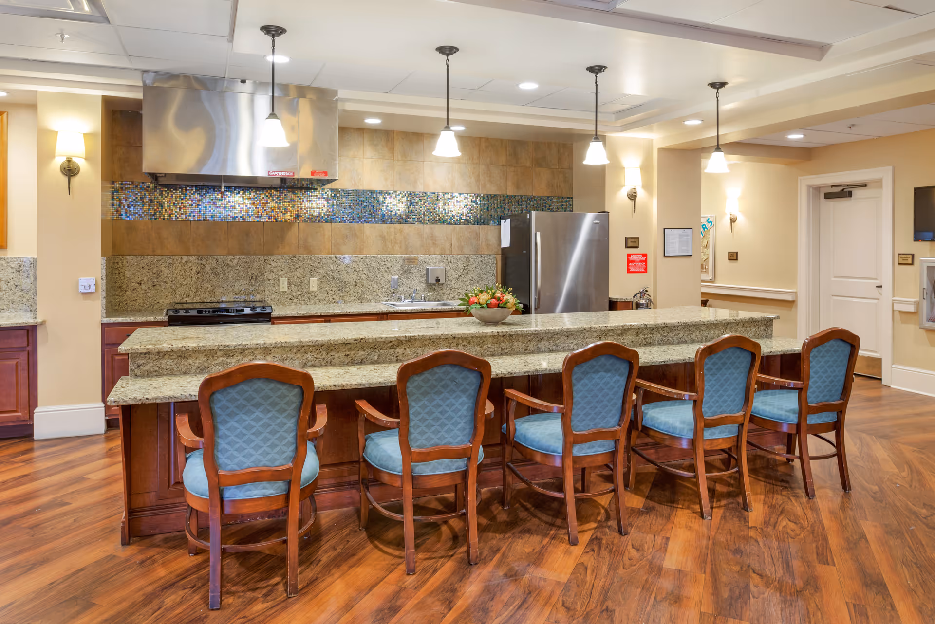 A modern kitchen area in a senior living facility featuring a long granite countertop with five wooden chairs upholstered in blue fabric. The kitchen has a stainless steel refrigerator, stove with a stainless steel range hood, a sink, and a colorful mosaic tile backsplash. The floor is wooden, and the area is well-lit with pendant lights and wall sconces.