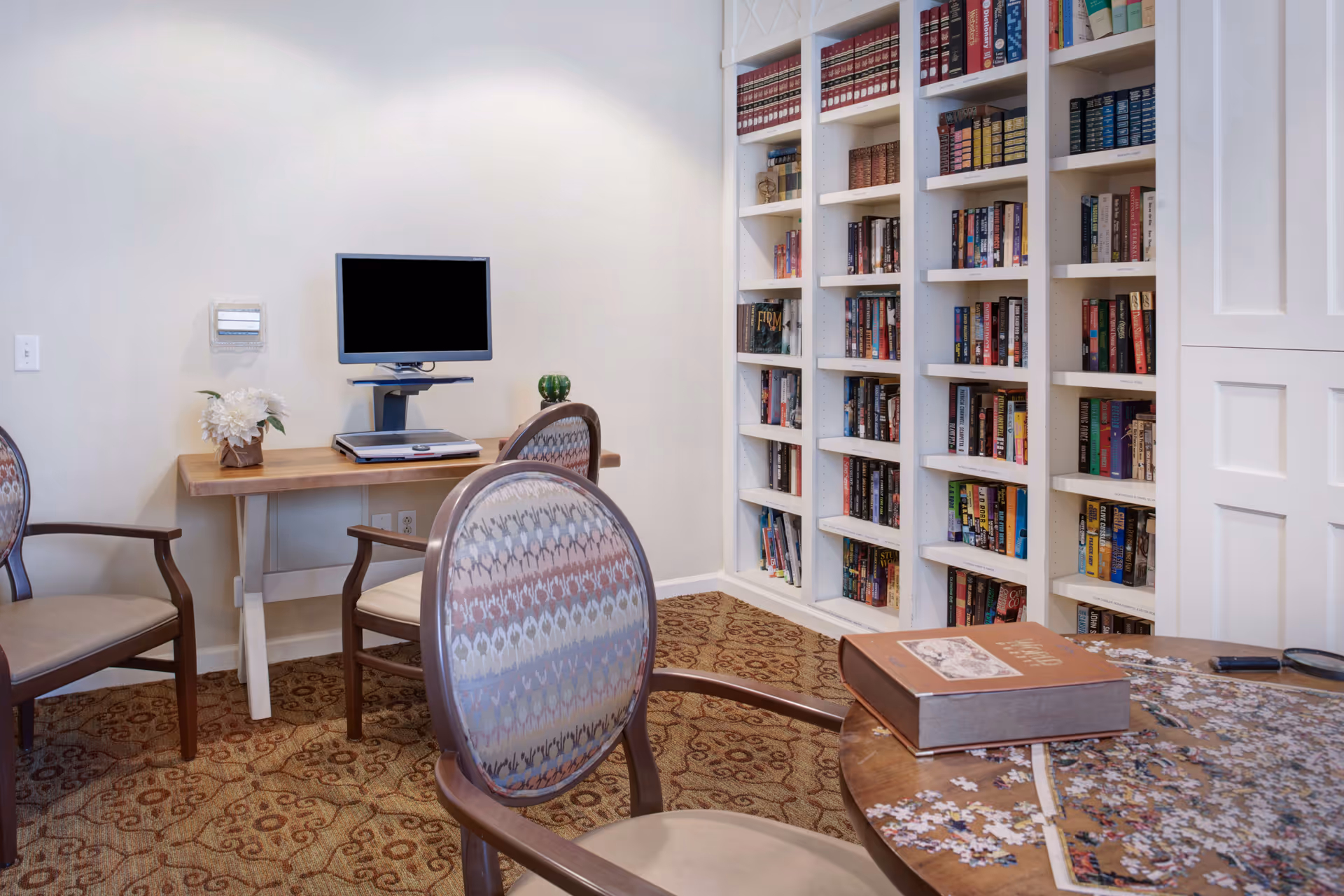 A cozy reading and activity room with a round table featuring a partially completed jigsaw puzzle and a large book. The room has patterned carpet, upholstered chairs, a wooden desk with a computer monitor, and a tall white bookshelf filled with books against the wall.