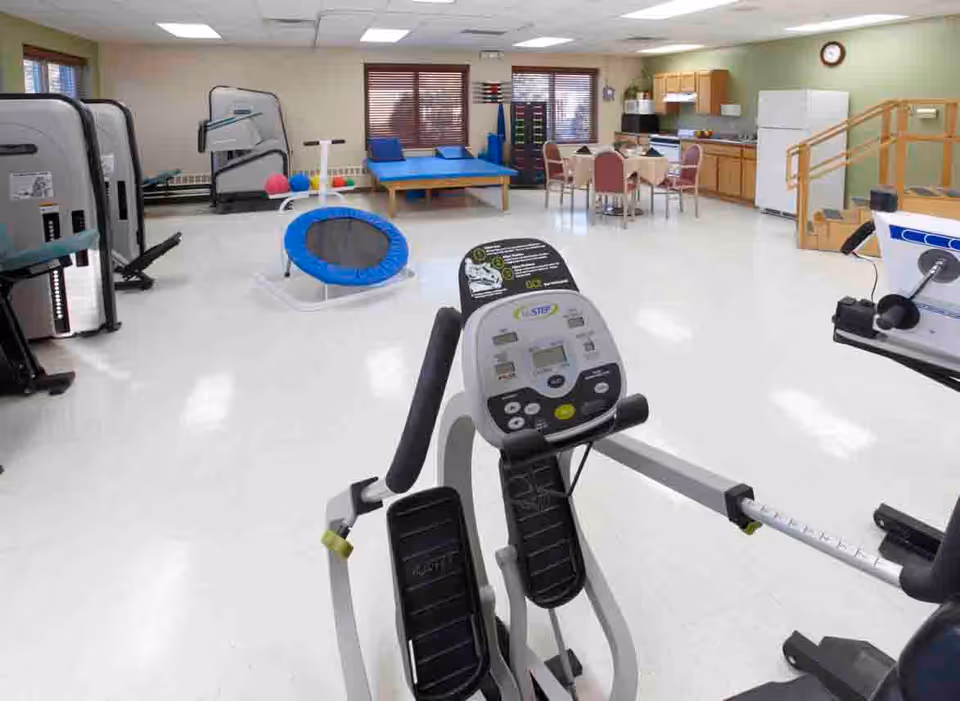 A spacious rehabilitation or physical therapy room with exercise equipment including a stationary bike, a small trampoline, therapy tables, and a table with chairs. The room has large windows with blinds, wooden cabinets, a refrigerator, and a clock on the wall.