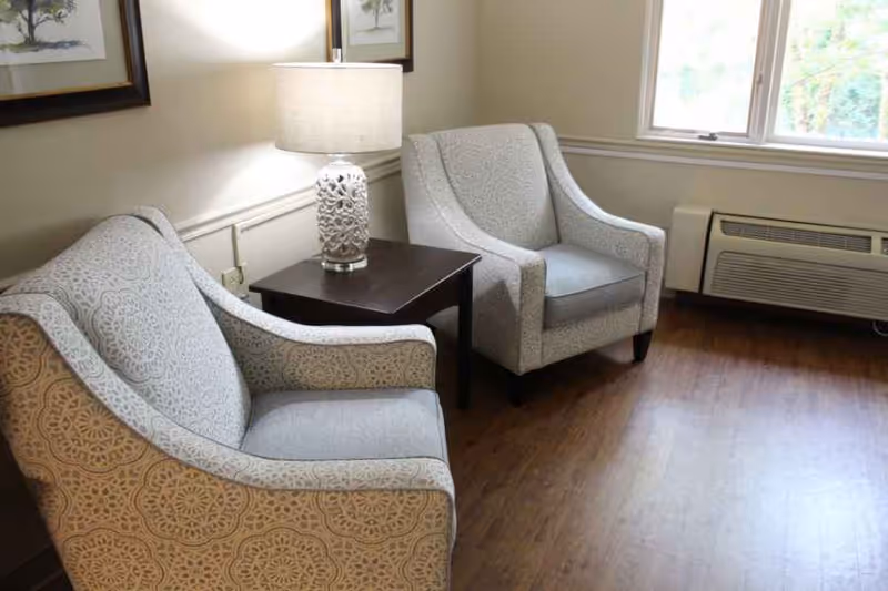 Two patterned armchairs flanking a side table with a lamp in a small seating area by a window and wall art.