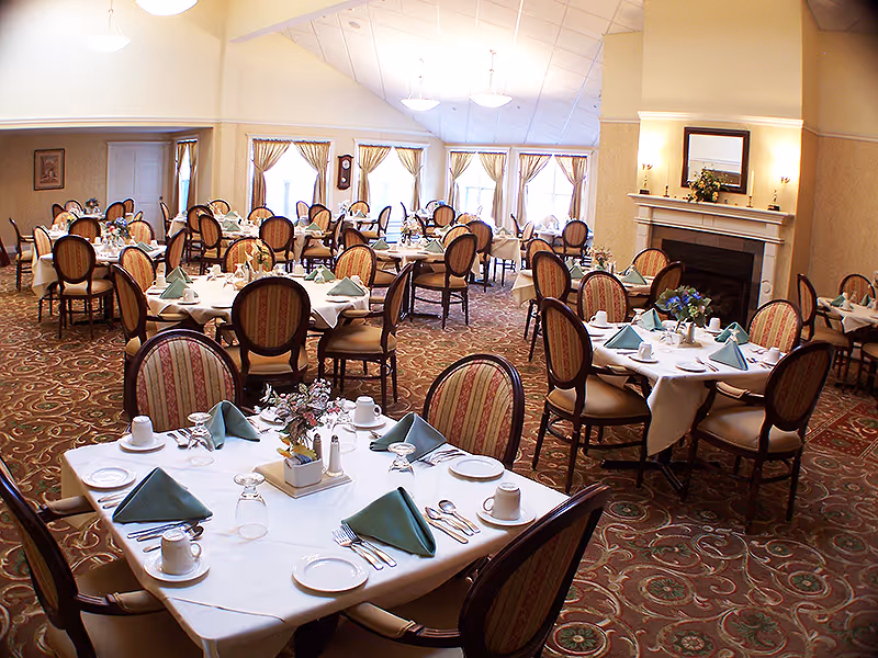 Bright formal dining room with multiple round tables set with white linens, folded napkins, and a fireplace along the far wall.
