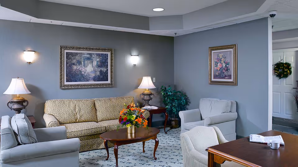 A cozy living room area with a patterned beige sofa, two light gray armchairs, a wooden coffee table with a colorful flower arrangement, two side tables with lamps, framed floral paintings on the walls, and a potted plant in the corner. The room has soft lighting and a carpeted floor with a subtle pattern.