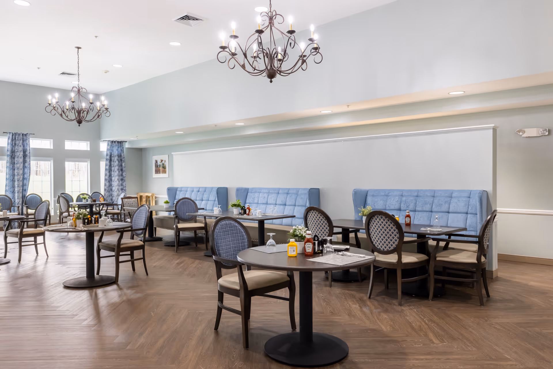 A bright and spacious dining room with multiple round tables and chairs. The room features light blue cushioned bench seating along the wall, dark wood chairs with patterned backs, and large windows with blue patterned curtains allowing natural light to fill the space. Two ornate chandeliers hang from the ceiling, and tables are set with condiments and glassware.