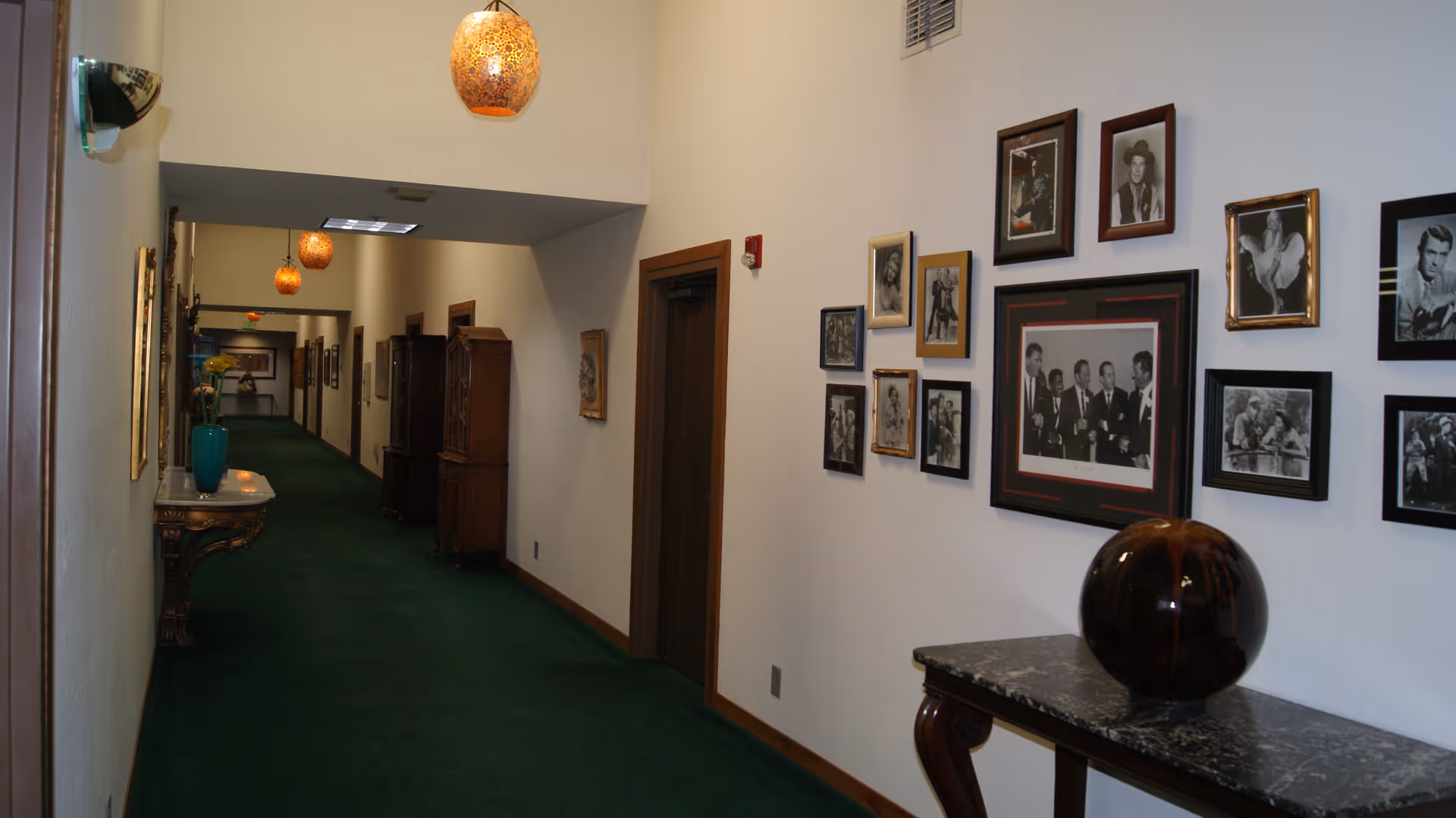 Long interior hallway with green carpet, framed photos on the right wall, decorative tables and hanging pendant lights.