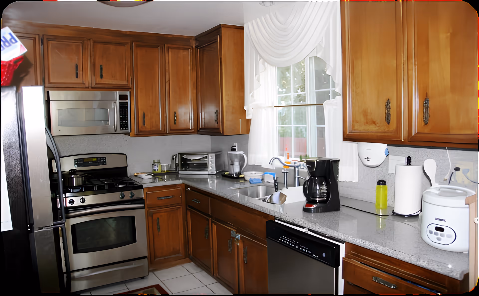 A kitchen with wooden cabinets, a stainless steel oven and microwave, a refrigerator, a toaster oven, a coffee maker, a rice cooker, and a sink under a window with white curtains.