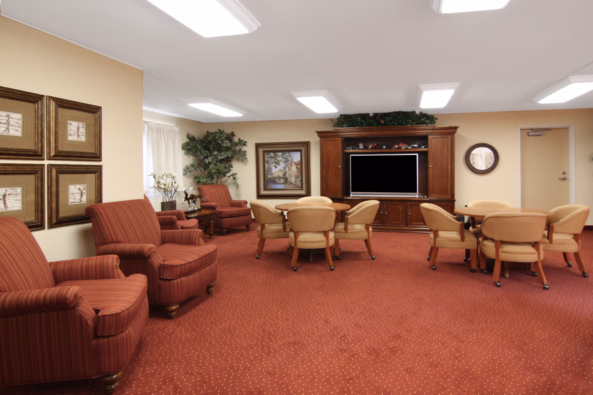 A spacious living room area with red patterned carpet, several upholstered armchairs, two round tables each surrounded by four beige chairs on casters, a large wooden entertainment center with a flat-screen TV, framed artwork on the walls, a clock, and potted plants.