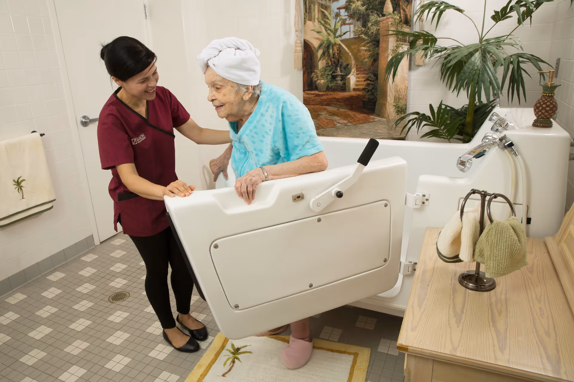 A caregiver assists an elderly woman with a towel wrapped around her head as she steps out of a walk-in bathtub in a bathroom. The bathroom has tiled floors and walls, a towel hanging on the wall, and a decorative plant on a wooden counter next to the tub.