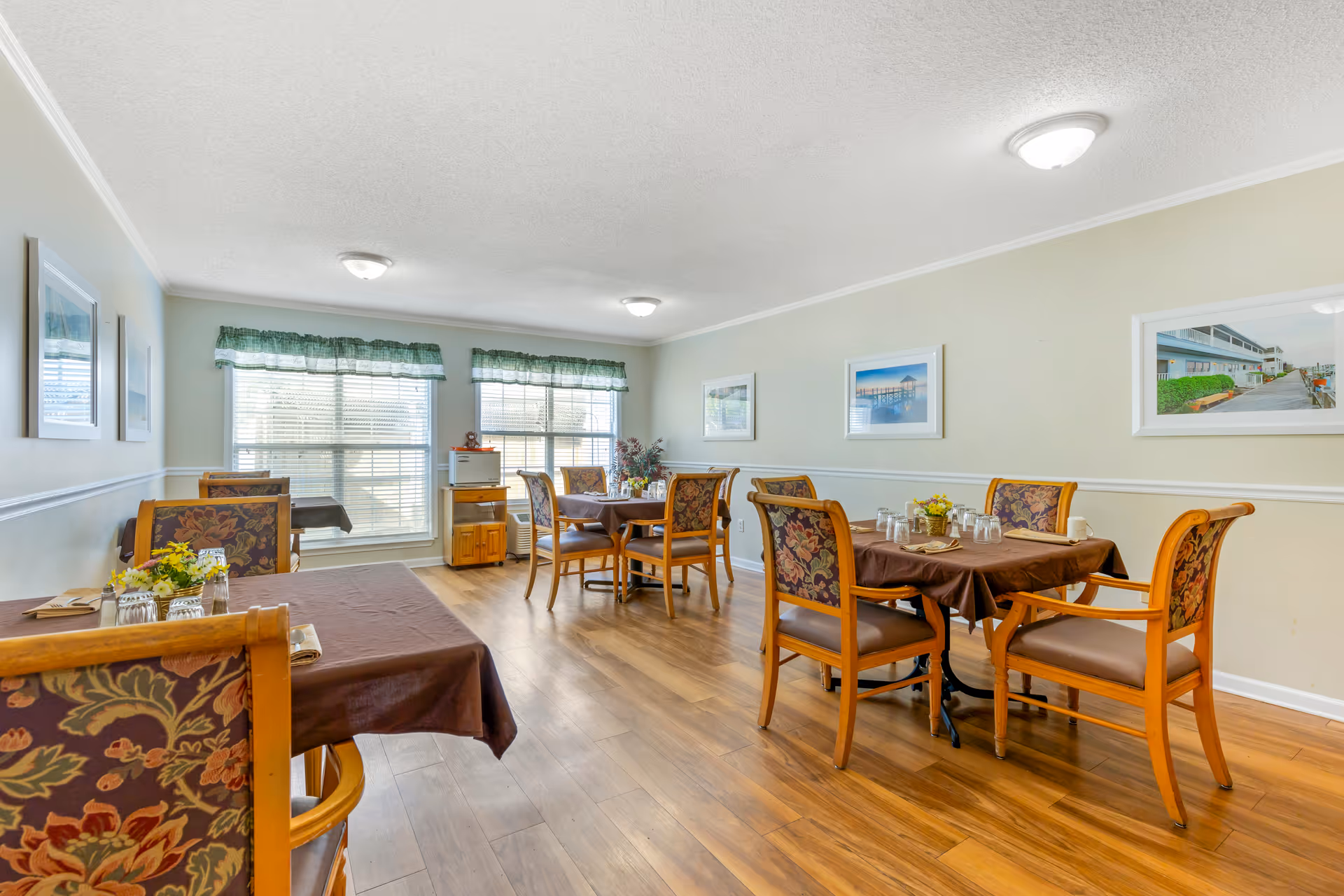 A dining room with wooden floors and several tables covered with brown tablecloths. Each table is surrounded by wooden chairs with floral upholstered backs and seats. The room has large windows with green valances letting in natural light. The walls are light-colored and decorated with framed pictures. There are ceiling lights providing additional illumination.