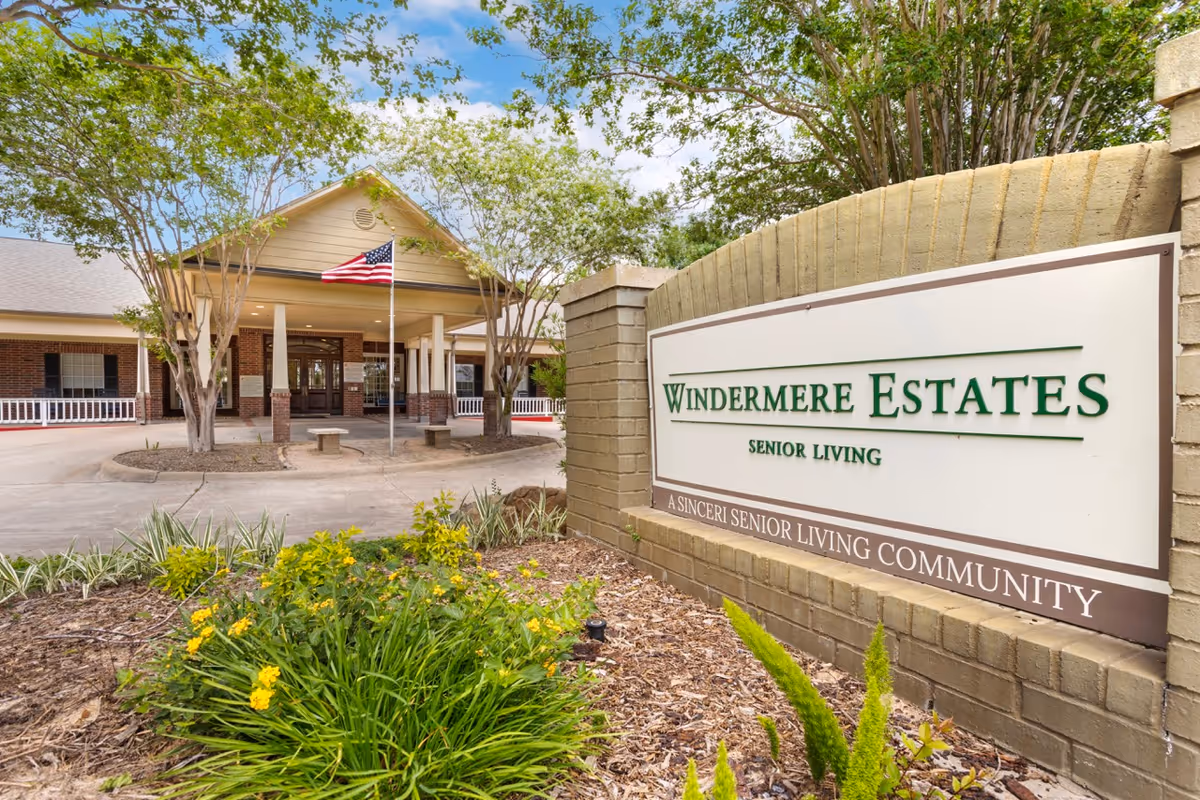 Entrance to Windermere Estates Senior Living community with a large sign in the foreground and a building with a covered driveway and American flag in the background, surrounded by trees and landscaping.